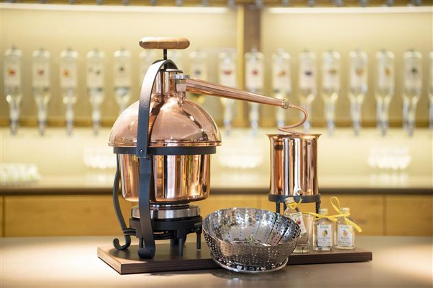 Een koperen distillatiekolf staat op een tafel. Op de achtergrond zijn verschillende flessen zichtbaar.