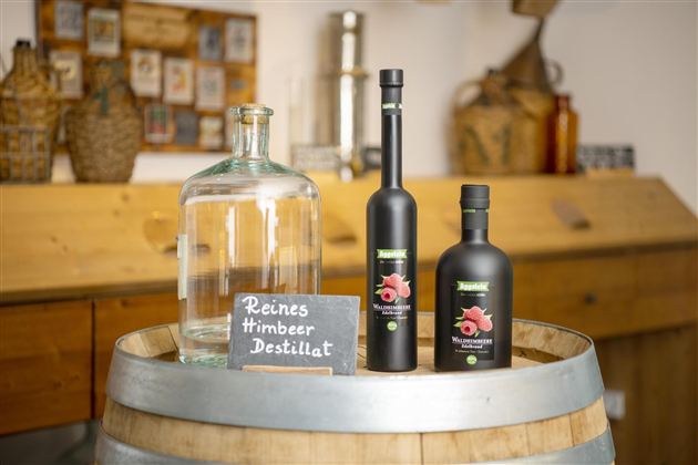 Een houten tafelblad met een grote glazen fles en twee flessen frambozen distillaat. Daarnaast staat een bord met de tekst "Puur Frambozen Distillaat".