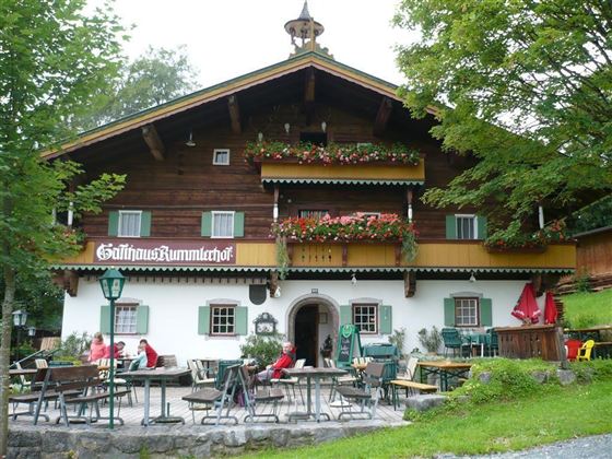 Ein traditionelles Holzhaus mit blühenden Balkonen und einer einladenden Terrasse. Gäste sitzen draußen und genießen die ruhige Umgebung.