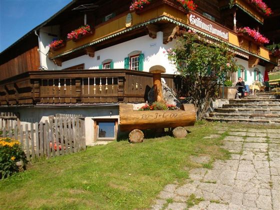 Ein traditionelles alpenländisches Haus mit Balkonen und Blumenkästen. Im Vordergrund befindet sich eine Holzbank und der Weg führt zu den Treppen.