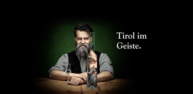 Een man met een baard houdt een lange houder vast, waaruit een gezicht verschijnt. Naast hem staat een fles, en de tekst zegt "Tirol in de geest".