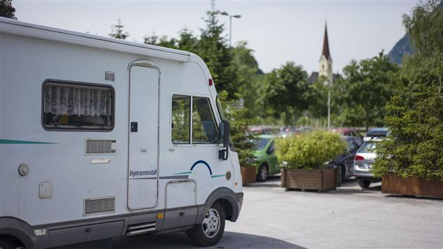 A motorhome in a parking lot, surrounded by trees and plants. In the background, a church steeple is visible.
