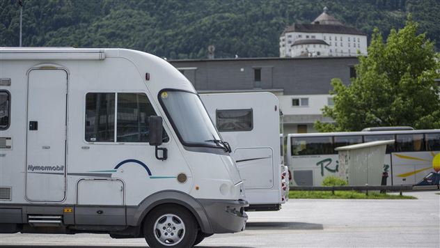 A motorhome is parked in a lot overlooking a mountainous landscape. In the background, a large building can be seen.