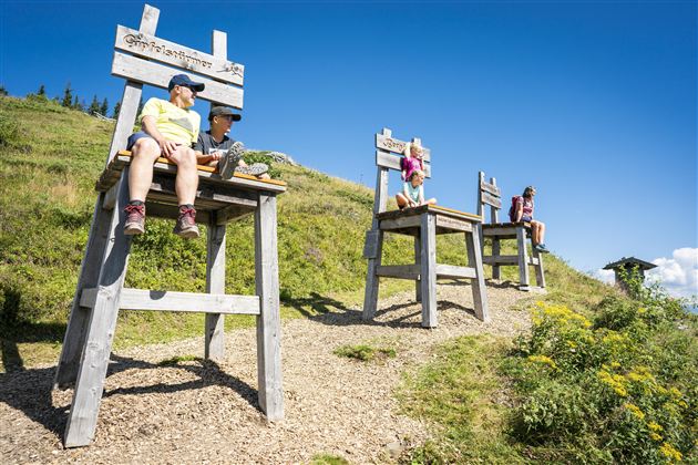 Large wooden chairs on a hilly terrain. People are sitting on them and enjoying the view in sunny weather.