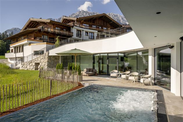 A modern house with large windows and an adjacent pool. In the background, green meadows and mountains can be seen.