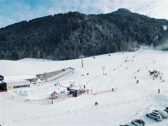 Sněhem pokrytá krajina se sjezdaři a lanovkou v pozadí. Nebe je částečně zatažené a hory jsou pokryté stromy.