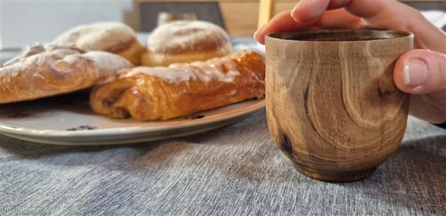 A handcrafted wooden cup is held by a hand. In the background, various pastries are visible on a plate.