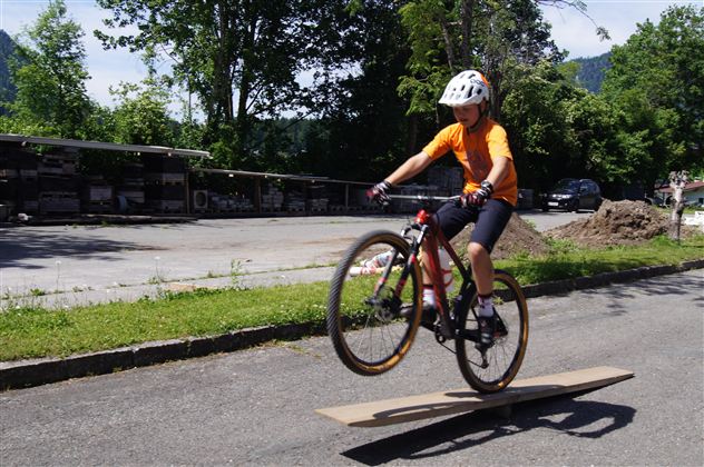 Ein Radfahrer springt über ein Holzbrett auf einer Straße. Er trägt einen Helm und ein orangefarbenes Shirt.