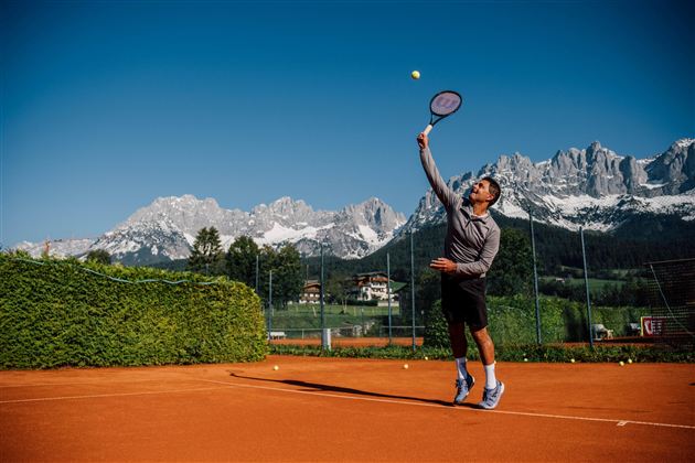 Ein Tennisspieler serviert auf einem Sandplatz. Im Hintergrund sind majestätische Berge zu sehen.