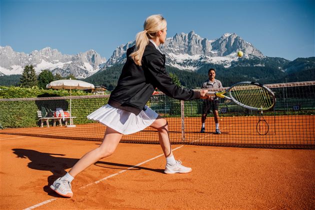 Eine Frau spielt Tennis auf einem Platz mit Blick auf beeindruckende Berge. Es ist sonnig und die Atmosphäre ist aktiv und sportlich.