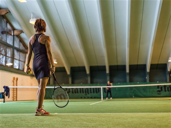 Eine Tennisspielerin steht auf dem Platz und blickt zum Netz. Der Innenbereich ist gut beleuchtet mit einer grünen Spielfläche.