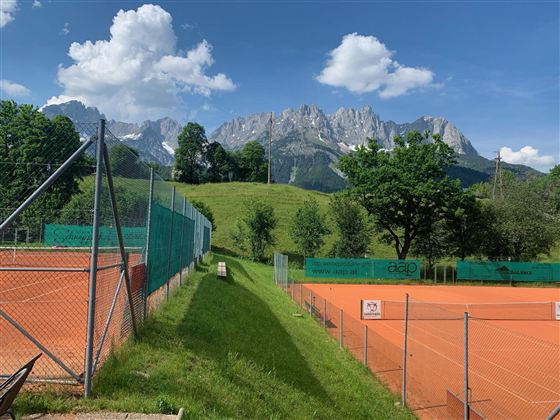 Eine Tennisplatzlandschaft mit orangefarbenem Belag und angrenzendem grünen Rasen. Im Hintergrund ragen beeindruckende Berge unter einem blauen Himmel hervor.