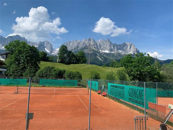 Ein Tennisplatz umgeben von grünen Wiesen und Bäumen. Im Hintergrund sind majestätische Berge und ein blauer Himmel mit weißen Wolken zu sehen.