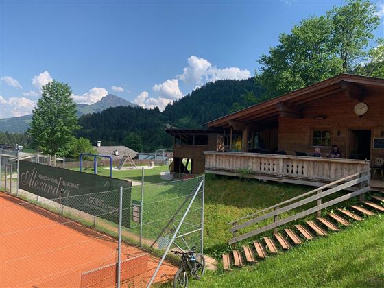 Eine gemütliche Hütte mit einer Veranda in einer malerischen Landschaft. Im Vordergrund ist ein Tennisplatz mit rotem Belag und umgebenden Bäumen zu sehen.
