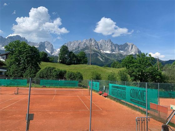 Ein Tennisplatz umgeben von grünen Bäumen und einer Berglandschaft. Die hohen Berge sind im Hintergrund mit blauem Himmel und weißen Wolken zu sehen.