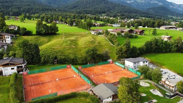 Eine Luftaufnahme von einem Tennisplatz mit rotem Sand, umgeben von grünen Wiesen und Bäumen. Im Hintergrund sind einige Häuser und Berge zu sehen.