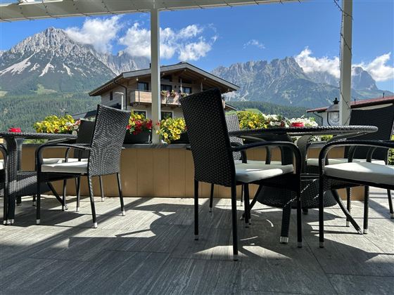 Eine gemütliche Terrasse mit schwarzen Stühlen und Tischen. Im Hintergrund sind majestätische Berge und ein blauer Himmel zu sehen.