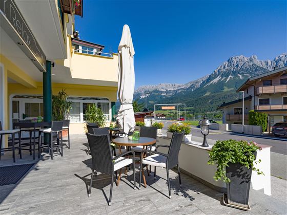 Ein gemütlicher Außenbereich mit Tischen und Stühlen. Im Hintergrund sind schöne Berge und ein strahlend blauer Himmel zu sehen.
