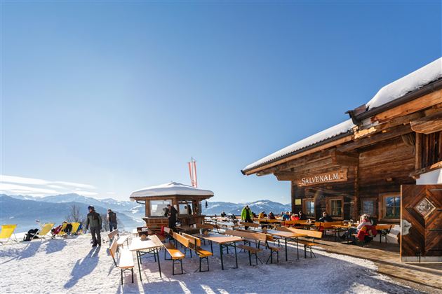 A cozy mountain restaurant with outdoor seating in the snow. The sky is blue and the surroundings are wintry and sunny.