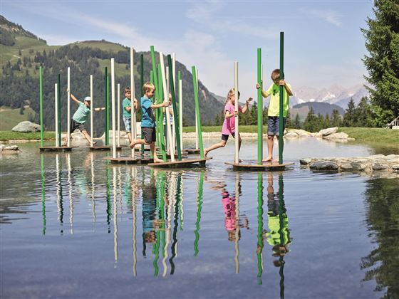 Vijf kinderen staan op platforms in het water en balanceren met kleurrijke stokken. Op de achtergrond zijn groene heuvels en een heldere lucht te zien.
