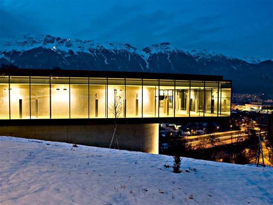 Ein modernes Gebäude mit großen Glasfenstern steht auf einem Hügel im Schnee. Im Hintergrund sind schneebedeckte Berge und ein Nachtblick auf die Stadt zu sehen.