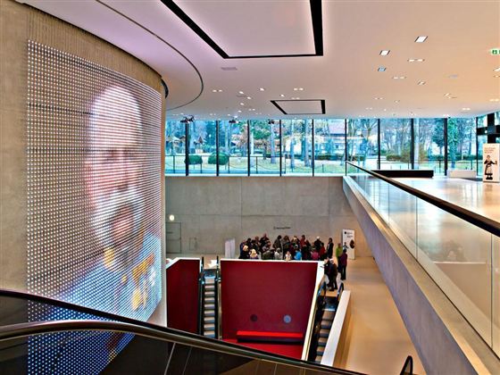 Ein modernes Foyer mit einer großen LED-Wand, die ein Bild zeigt. Besucher stehen in einer Warteschlange im unteren Bereich.
