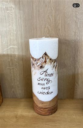 Eine dekorative Kerze mit einem schönen Muster und der Aufschrift "Amoi seng ma uns wieder". Sie steht auf einem Holzuntergrund.