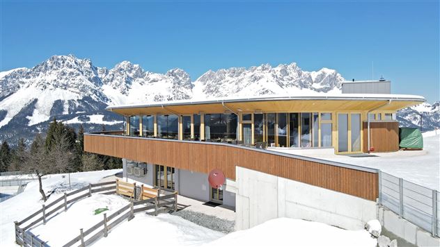Een modern gebouw in de bergen, omgeven door sneeuwbedekte landschappen. Op de achtergrond zijn indrukwekkende bergtoppen onder een heldere blauwe lucht te zien.