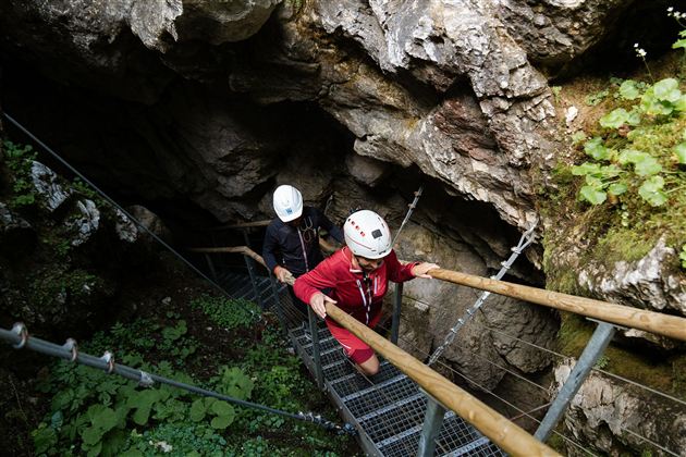 Two people are descending a staircase into a cave. They are wearing helmets and are surrounded by rocks and plants.