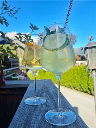 Twee grote glazen met verfrissende cocktails staan op een houten reling. Op de achtergrond straalt de blauwe lucht en het groene landschap.