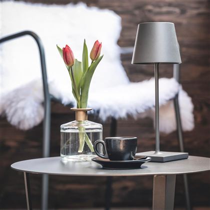 Ein stilvoller Tisch mit einer Tasse Kaffee und einer Glasvase mit frischen Tulpen. Im Hintergrund steht eine moderne Lampe und ein gemütlicher Stuhl.