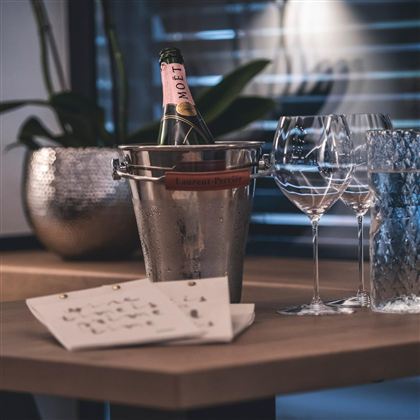 Ein eleganter Tisch mit einem Champagnerkübel, der eine Flasche Moët enthält. Daneben stehen zwei Weingläser und eine schöne Karte.