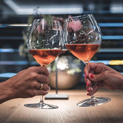 Zwei Gläser mit Roséwein werden angestoßen. Im Hintergrund sind verschwommene Blumen und sanftes Licht zu sehen.