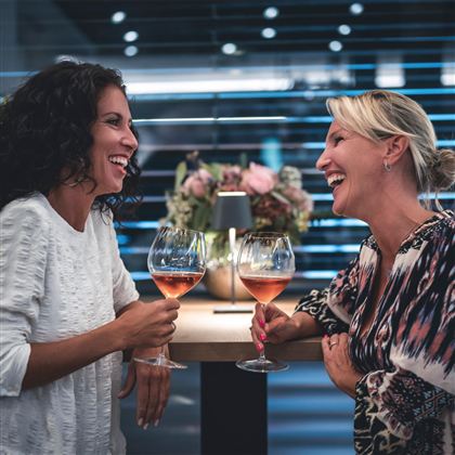 Zwei Frauen lachen und stoßen mit Weingläsern an. Im Hintergrund sind Blumen und eine moderne Umgebung zu sehen.