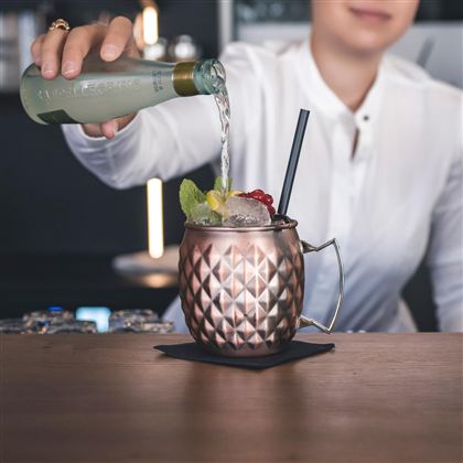 Ein Barkeeper gießt ein Getränk in einen eleganten, kupfernen Becher. Der Becher ist mit Eis, Limetten und roten Beeren dekoriert.