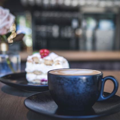 Eine Tasse Kaffee auf einem Teller neben einem Stück Kuchen. Im Hintergrund sind stilvolle Dekorationen zu sehen.