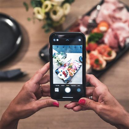 Eine Person hält ein Smartphone, um ein Foto von einer schönen platzierten Essensauswahl zu machen. Im Hintergrund sind frische Zutaten und eine dekorative Anordnung sichtbar.