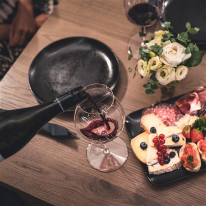 Ein Glas Rotwein wird eingeschenkt, während ein rustikales Essen auf einem Tisch präsentiert wird. Dazu gehören Käse, Wurst, Obst und frische Blumen.