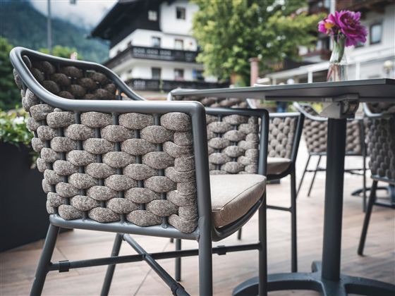 Ein moderner Stuhl aus geflochtenem Material steht an einem Tisch. Im Hintergrund sind grüne Bäume und Gebäude zu sehen.