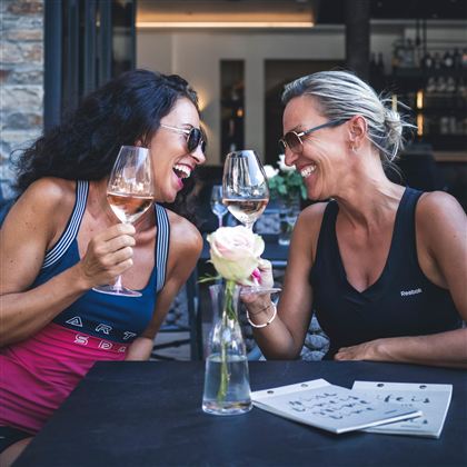 Zwei Frauen lachen und stoßen mit Gläsern an, während sie am Tisch sitzen. Auf dem Tisch steht eine Rose und einige Menükarten.