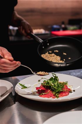 Ein Koch präsentiert einen Teller mit dünn geschnittenem Fleisch, garniert mit frischen Kräutern. Im Hintergrund wird eine Zutat aus einer Pfanne auf den Teller gestreut.