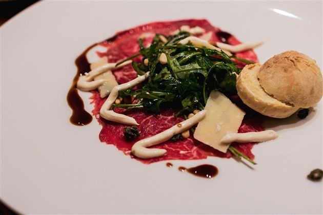 Ein hübsches Gericht mit dünn geschnittenem Fleisch, frischem Blattspinat und einem kleinen Brötchen. Die Anrichtung ist geschmackvoll mit einer cremigen Sauce und Balsamico-Reduktion.