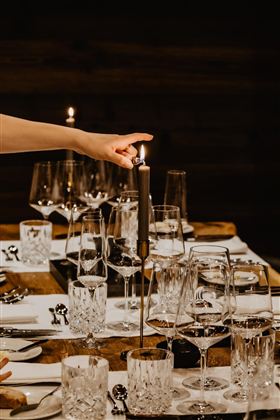 Ein eleganter Esstisch mit Gläsern und Besteck. Eine Hand zündet eine Kerze an und sorgt für gemütliches Licht.