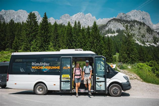 A hiking bus stands in nature, surrounded by mountains and forests. Two people get out, ready for an adventure.