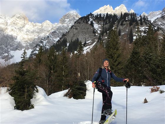 Eine Frau beim Schneeschuhwandern in einer verschneiten Berglandschaft. Im Hintergrund sind majestätische Berge und Nadelbäume zu sehen.