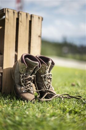 Een paar wandelschoenen staat op het gras naast een houten kist. De lucht is blauw en er zijn enkele wolken te zien.