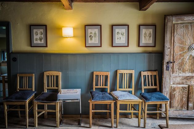 A cozy room with a row of wooden chairs and a rustic door. Pictures in frames hang on the wall, and a lamp illuminates the area.
