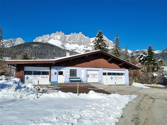 Een gezellig huis in de sneeuw met garages en een blauwe lucht. Op de achtergrond zijn de bergen zichtbaar.