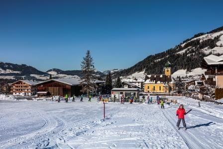 A snowy winter landscape with skiers and a picturesque backdrop. In the background, mountains and traditional alpine buildings can be seen.