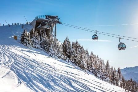 A ski lift station in the mountains, surrounded by snow-covered trees. In the background, gondolas glide over the snowy slope.